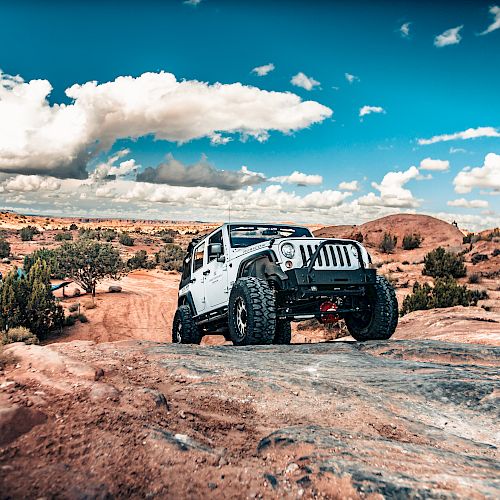 A white off-road vehicle navigating rocky terrain in a desert landscape with a clear blue sky and scattered clouds, with a campsite visible in the background.