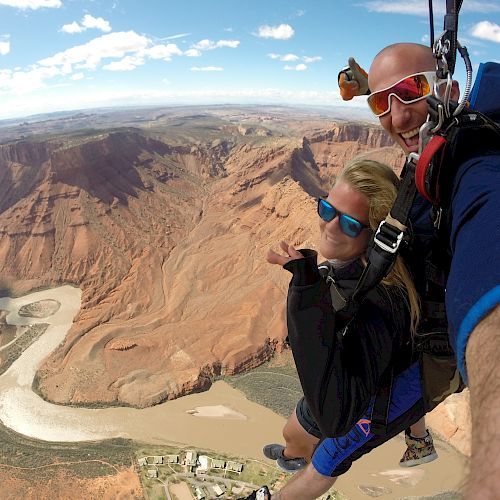 Two people are tandem skydiving over a scenic canyon landscape with a river in view, taking a selfie during their descent.