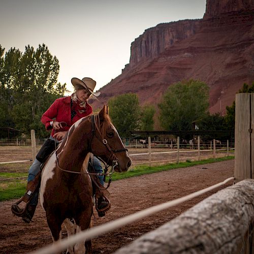 A person in a red shirt and cowboy hat rides a horse along a fenced dirt path with scenic cliffs and trees in the background.