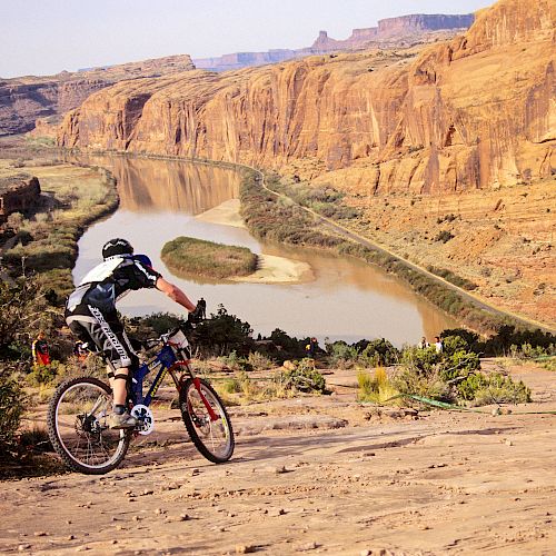 A person is mountain biking on a rocky path overlooking a winding river in a canyon landscape with rugged, orange cliffs in the background.