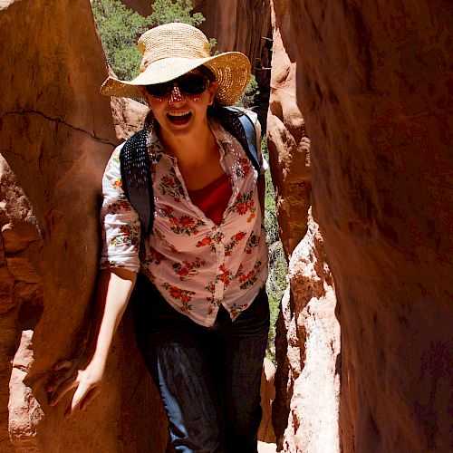 A person wearing a hat and sunglasses is standing between narrow rock formations, smiling and appearing to enjoy what seems to be a hiking adventure.