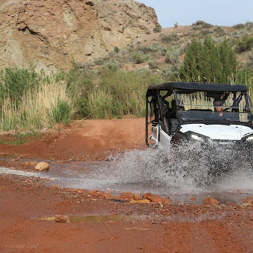A white off-road vehicle splashes through a muddy water crossing on a red dirt trail surrounded by rocky terrain and greenery.