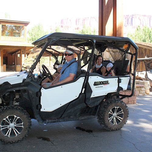 A group of people in a side-by-side ATV parked under a canopy with wooden buildings in the background.