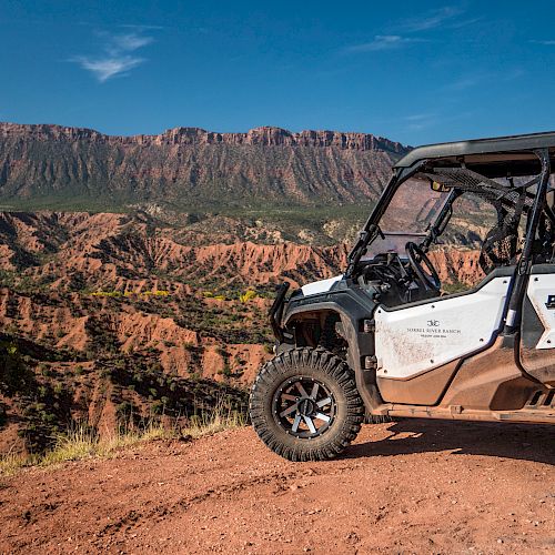 An off-road vehicle is parked on a dirt path overlooking a scenic canyon with rugged hills and clear blue skies in the background.
