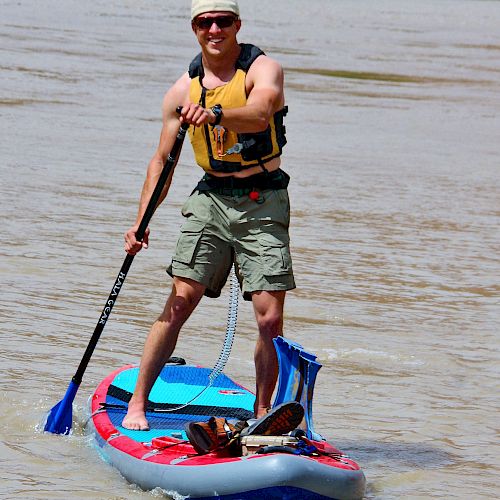 A person is paddleboarding on a river, wearing a life vest, hat, sunglasses, and shorts, holding a paddle with one foot forward.