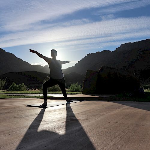 A person is silhouetted while performing a yoga pose outdoors with mountains and a setting sun in the background.