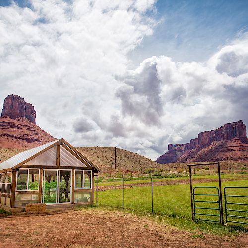 A small greenhouse stands in an open field with a fence and gate, set against a backdrop of red rock formations and a partly cloudy sky.