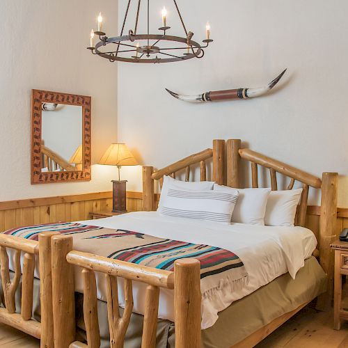 A cozy rustic bedroom with a wooden bed, decorative horn above, two lamps, a mirror, and a window reflecting another room, creating a warm atmosphere.