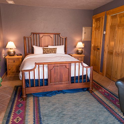 A cozy bedroom with a wooden bed frame, bedside tables with lamps, potted plant, colorful rug, and a leather chair.