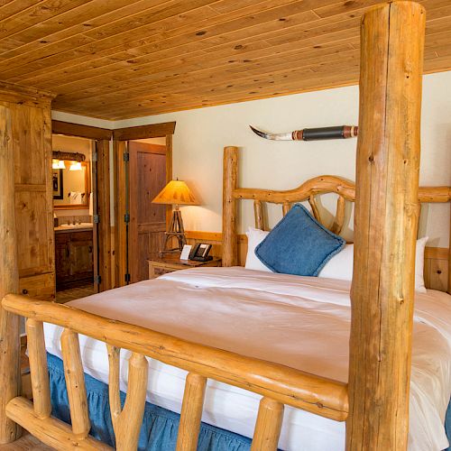 The image shows a rustic bedroom with wooden furniture, a large bed, two bedside tables with lamps, and a wooden ceiling.