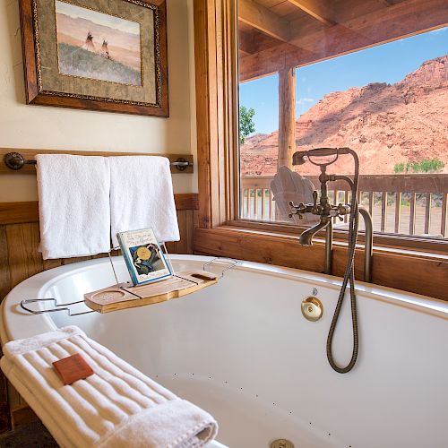 A bathtub with a towel, soap, and tablet holder overlooking a scenic desert landscape through a large window in a rustic bathroom.