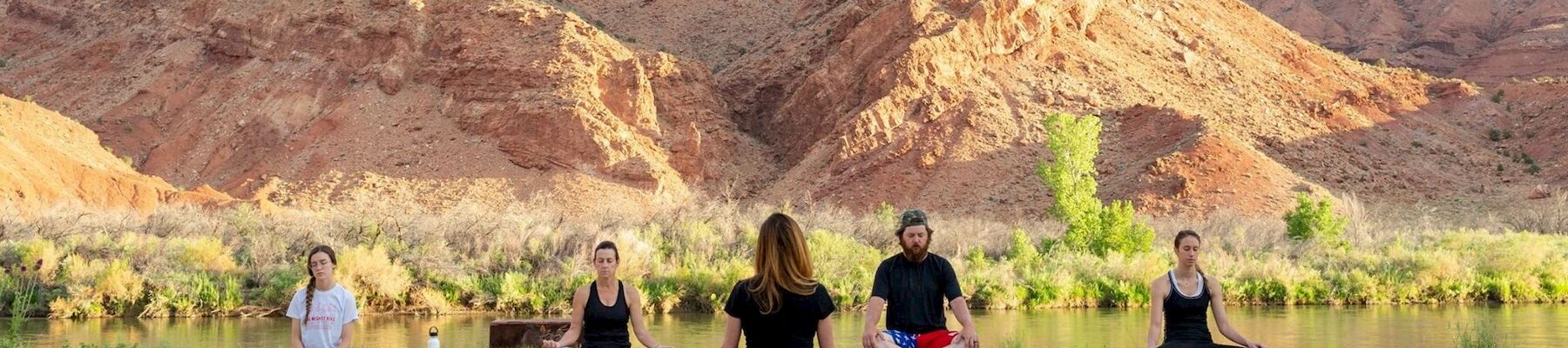 People are practicing yoga outdoors on a wooden platform with a scenic backdrop of rugged mountains and a river.