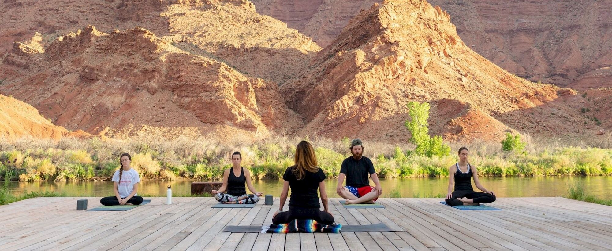 People are practicing yoga outdoors on a wooden platform with a scenic backdrop of rugged mountains and a river.