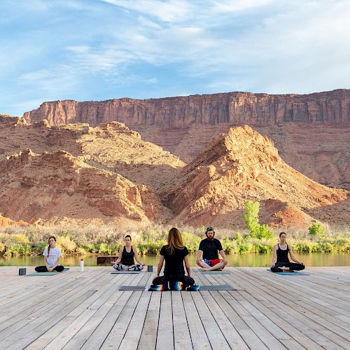 A group of people practicing yoga on a wooden platform with stunning canyon scenery in the background under a blue sky, creating a serene atmosphere.