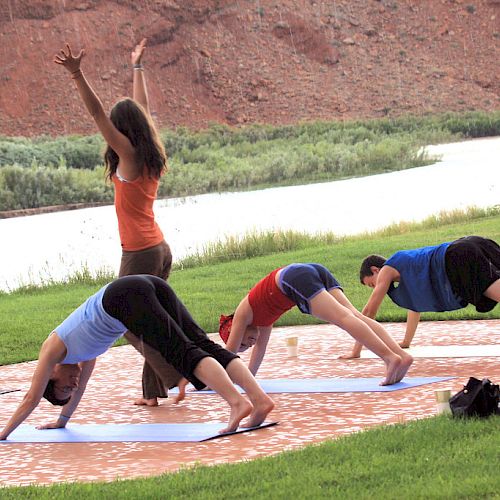 A yoga instructor is leading three individuals in an outdoor yoga session by a river with a scenic background.