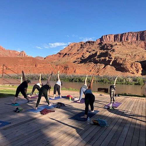 People are practicing outdoor yoga on a wooden platform, with scenic red rock cliffs and a river in the background, under a clear blue sky.