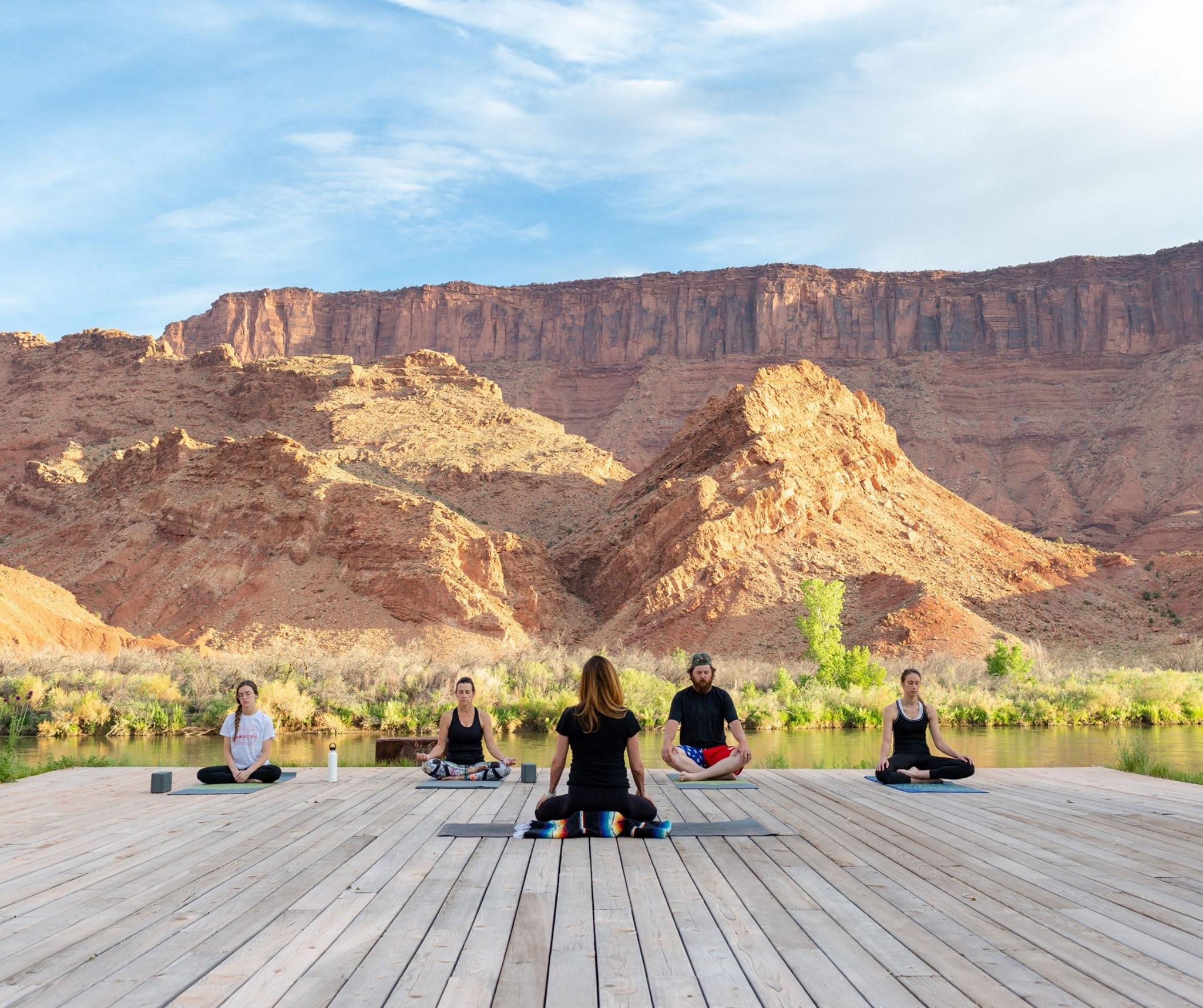 A group of people practicing yoga on a wooden deck overlooking a river and mountains.