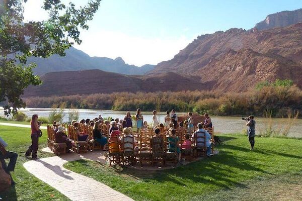 A group of people are gathered outdoors by a river with mountains in the background, sitting on chairs arranged in a circle.