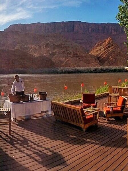 An outdoor dining setup on a deck by a river with mountains in the background, featuring wooden furniture, a grill, and a person standing by a table.