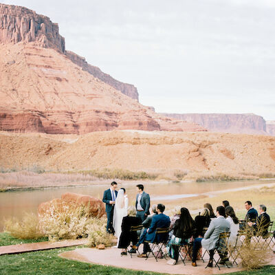 This image depicts a small outdoor wedding ceremony taking place in a picturesque desert setting near a river, with red rock formations in the background.