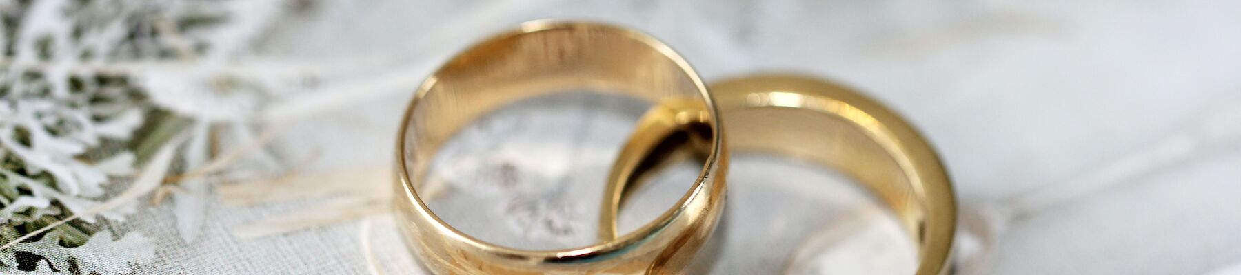 The image shows two gold wedding rings placed on a white decorative surface with greenery in the background.