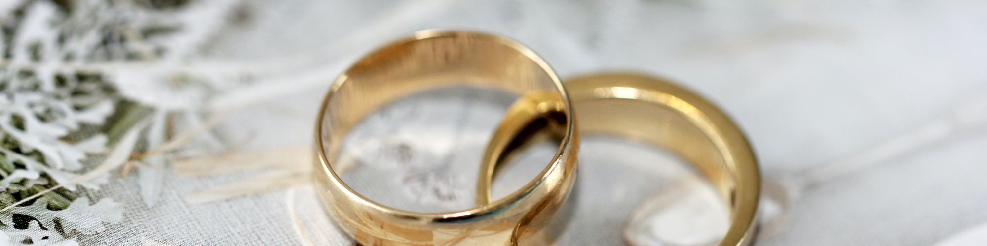 The image shows two gold wedding rings placed on a white decorative surface with greenery in the background.