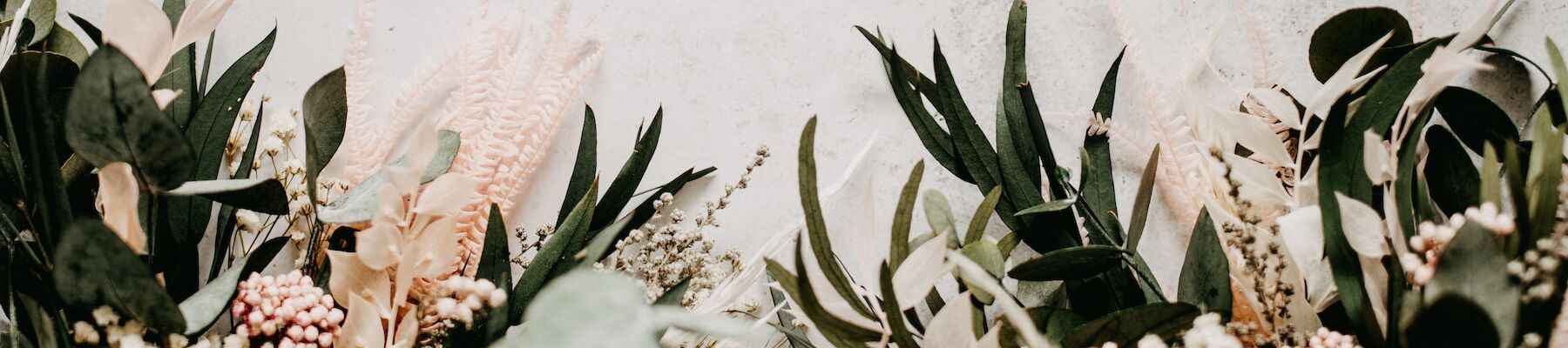The image shows a floral arrangement with a variety of leaves and small flowers at the bottom against a textured, light-colored background.