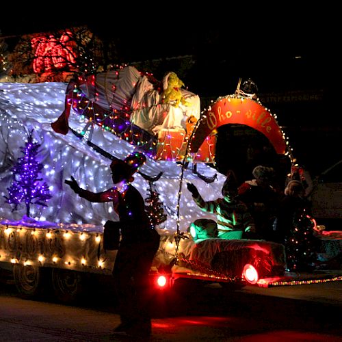 A decorated float with Christmas lights and people dressed festively is moving in a parade at night.
