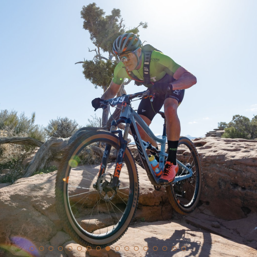 A person is mountain biking over rocky terrain under a clear sky, wearing a helmet, sunglasses, and cycling gear, with bright sunlight in the background.