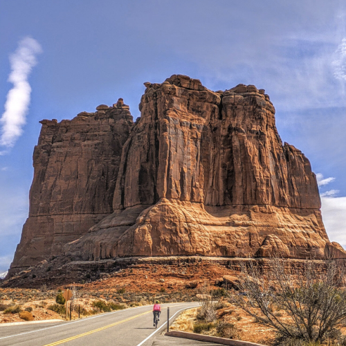 A towering red rock formation stands beside a paved road under a blue sky with scattered clouds. A person walks towards the formation.