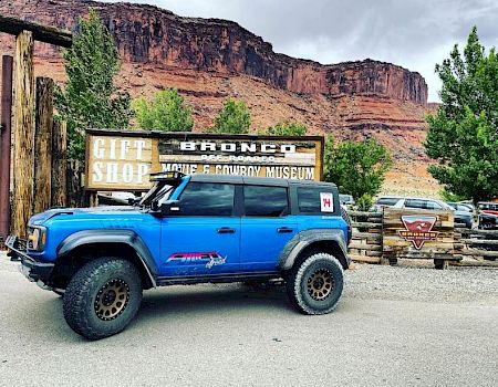 A blue off-road vehicle is parked near a sign for a gift shop and a movie car museum, with rocky cliffs and trees in the background.