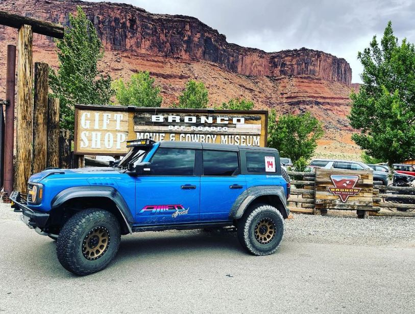 A blue off-road vehicle is parked near a sign for a gift shop and a movie car museum, with rocky cliffs and trees in the background.