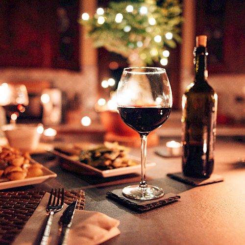A table set for a romantic dinner with a glass of red wine, wine bottle, plates of food, and lit candles, creating a cozy, intimate atmosphere.