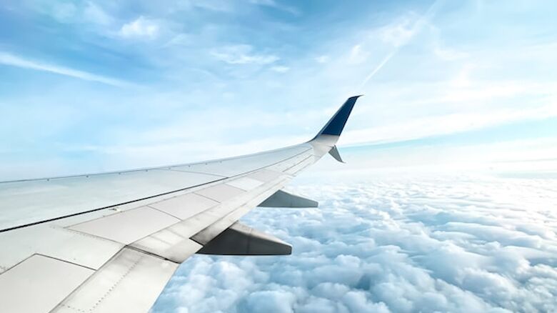The image shows the wing of an airplane flying above fluffy clouds with a bright blue sky in the background.