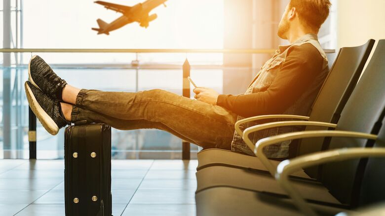 A person is sitting in an airport lounge with their feet on a suitcase, looking at a plane taking off through the window.