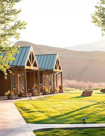Cozy cabins with blue roofs, surrounded by lush greenery and trees, with a mountain backdrop under warm sunlight.