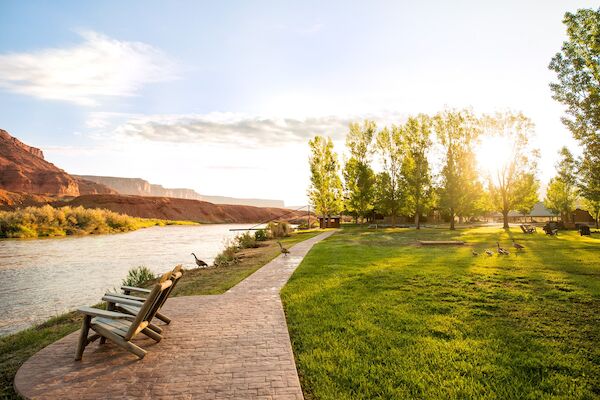 A riverside park at sunset with trees, grass, and wooden chairs. A path runs along, with geese near the water.