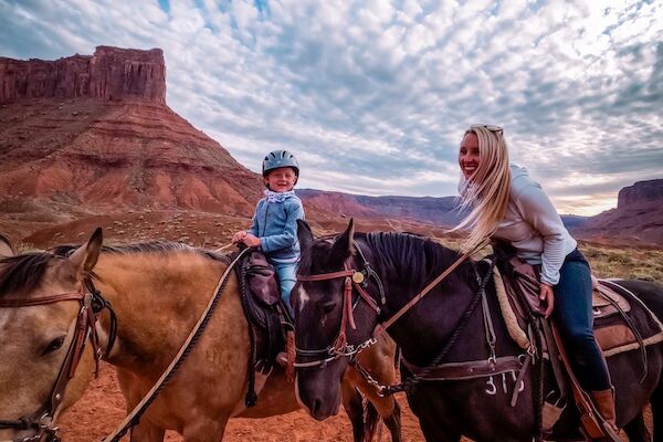 A child and an adult are horseback riding in a desert landscape with stunning rock formations and a sky filled with clouds.