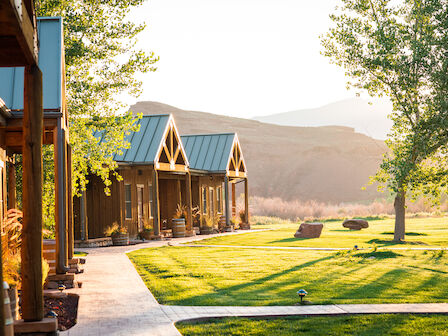 The image features a serene outdoor scene with wooden cabins, a manicured lawn, pathway, and trees in a mountainous area, bathed in sunlight.