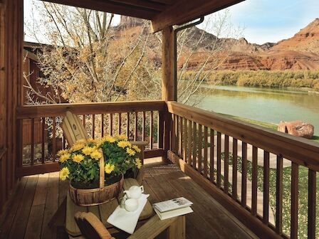 Wooden porch with a table, yellow flowers, and a lake view with mountains in the background. A book and cup are on the table.