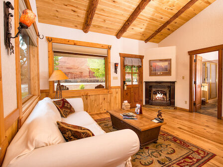 A cozy wooden living room with a white sofa, coffee table, fireplace, and large windows with a scenic view. The door leads to another room.