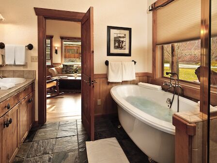 The image shows a cozy bathroom with a soaking tub, wooden cabinetry, and a view into a comfortable living room with rustic decor, ending the sentence.