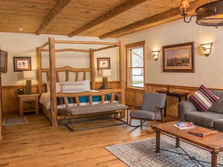 A cozy cabin-style room with a wooden post bed, sofa, armchair, wooden furniture, framed artwork, and natural light from the windows.