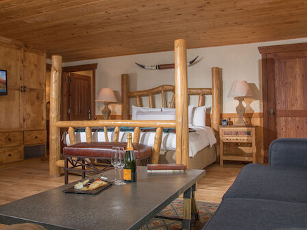 The image shows a rustic bedroom with wooden walls, a large log bed, a sofa, and a coffee table with wine and cheese. There's a mounted TV.