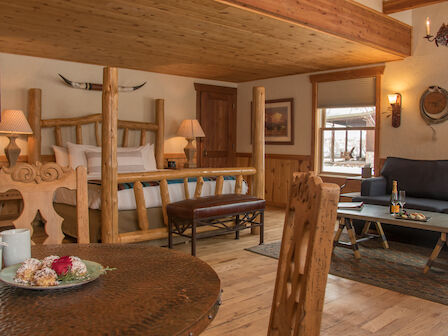 A cozy, rustic bedroom and living area with wooden decor, featuring a bed, couch, coffee table, and a small dining area with tea and snacks.