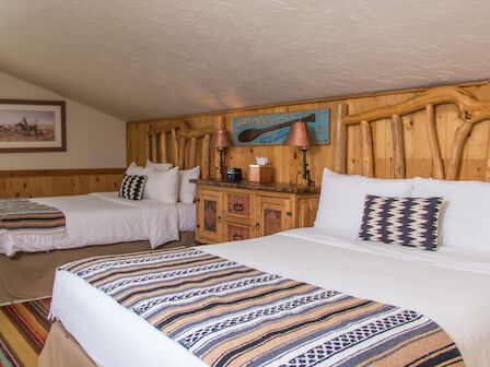 The image shows a cozy hotel room with two beds, rustic wooden headboards, patterned bed runners, and a window offering natural light and a view.