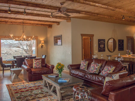A cozy, rustic living room with wooden ceilings, leather sofas, a colorful rug, a dining table by the window, and a kitchen corner with barstools.