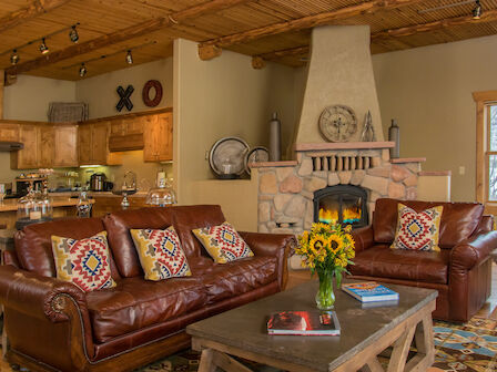 A cozy living room with leather sofas, patterned pillows, a stone fireplace, and a wooden ceiling opens to a kitchen area with rustic decor.