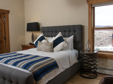 A cozy bedroom with a gray tufted bed, blue and white blanket, nightstand, lamp, and window with a scenic outside view in the background.