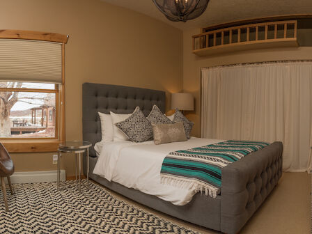 A cozy bedroom with a large bed adorned with pillows and a striped throw blanket, a chair by the window, and a decorative rug on the floor.